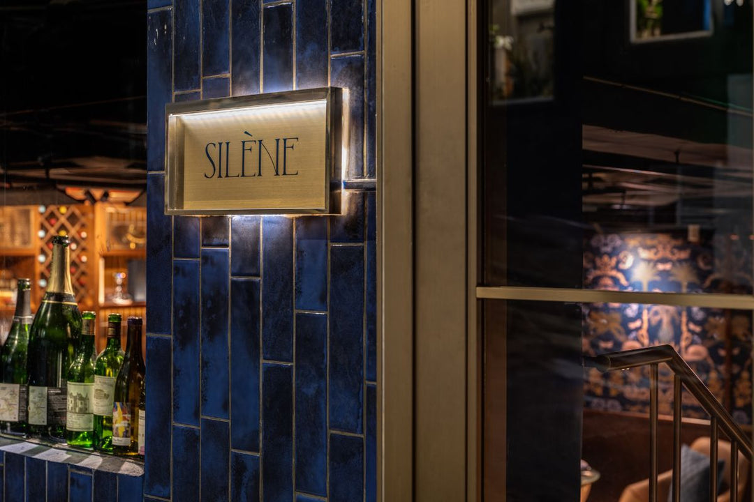 A sign reading "SILÈNE" is mounted on a blue-tiled wall next to a glass window; bottles are visible on a counter inside.